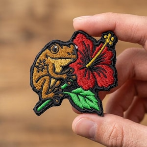May include: Embroidered patch featuring a brown frog with yellow accents, perched next to a red hibiscus flower with a yellow stamen and green leaves. The patch has a black border.