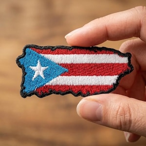 May include: Embroidered patch in the shape of Puerto Rico's island, featuring the flag's design. The patch has a blue triangle with a white star, and red and white stripes. The patch is held by a hand.