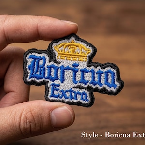 May include: Embroidered patch with the text "Boricua Extra" in blue and white, with a gold crown design. The patch has a black border and is held in a hand. The text "Style - Boricua Extra" is at the bottom.