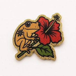 May include: A whimsical frog design with a red hibiscus flower and green leaves. The frog is tan with a black sunburst design on its side. The edges are outlined in gold glitter.