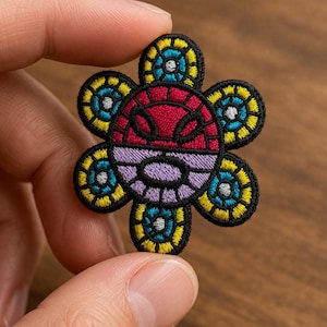May include: Embroidered flower patch with a black outline, featuring a red, purple, yellow, and blue design. The patch is held between fingers, showcasing the intricate details of the colorful floral pattern. This decorative patch can be applied to clothing or accessories.