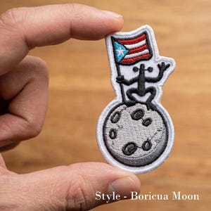 May include: An embroidered patch featuring a black Coqui frog sitting on a grey moon, holding a flag with red, white, and blue stripes and a white star on a blue triangle. The patch has a white border and the text Style - Boricua Moon.