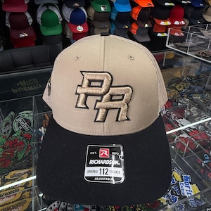 May include: A tan and black Richardson 112 trucker hat with the letters "PR" embroidered in a gold color. The hat has a mesh back and an adjustable strap. The hat is displayed in a store with other hats.