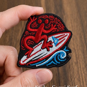 May include: An embroidered patch featuring a red coqui frog surfing on a white surfboard with a blue and white star emblem. The design includes blue wave patterns at the bottom and floral accents in red on a black background. Text reads Style - Coqui Surf.