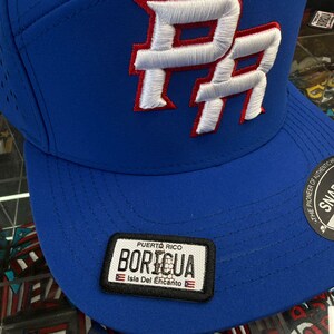 May include: A blue baseball cap with a white and red embroidered "PR" logo. The cap features a patch with the text "Puerto Rico Boricua Isla Del Encanto". The hat has a curved brim and a snap closure.