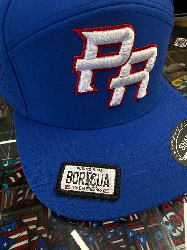 May include: A blue baseball cap with a white and red embroidered "PR" logo. The cap features a patch with the text "Puerto Rico Boricua Isla Del Encanto". The hat has a curved brim and a snap closure.