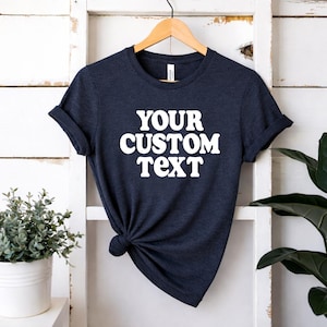 May include: A dark navy blue short-sleeve t-shirt featuring the white text "YOUR CUSTOM TEXT" printed in a bold, rounded font on the chest. The shirt has a crew neckline and is displayed on a wooden hanger with the fabric knotted at the side.