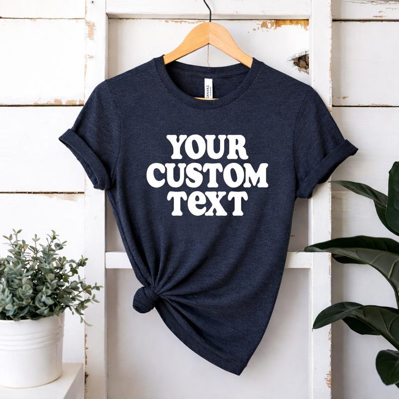May include: A dark navy blue short-sleeve t-shirt featuring the white text "YOUR CUSTOM TEXT" printed in a bold, rounded font on the chest. The shirt has a crew neckline and is displayed on a wooden hanger with the fabric knotted at the side.