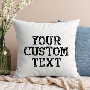 May include: A square white throw pillow featuring the black text YOUR CUSTOM TEXT centered on the front. The pillow has a smooth fabric finish and is positioned in front of a blue textured pillow and a clear glass vase with white flowers.