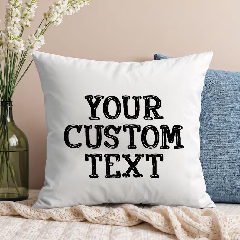 May include: A square white throw pillow featuring the black text YOUR CUSTOM TEXT centered on the front. The pillow has a smooth fabric finish and is positioned in front of a blue textured pillow and a clear glass vase with white flowers.