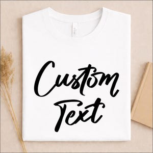 May include: A white short-sleeved t-shirt featuring the words Custom Text printed in a bold black script font across the center of the chest. The garment has a classic crew neckline and a smooth, clean finish on the front surface.