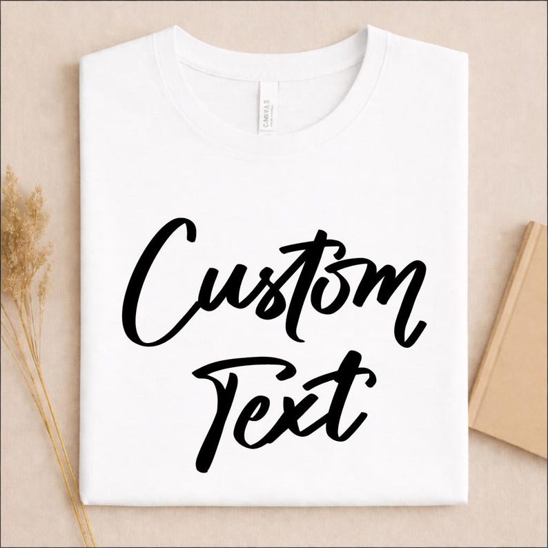 May include: A white short-sleeved t-shirt featuring the words Custom Text printed in a bold black script font across the center of the chest. The garment has a classic crew neckline and a smooth, clean finish on the front surface.
