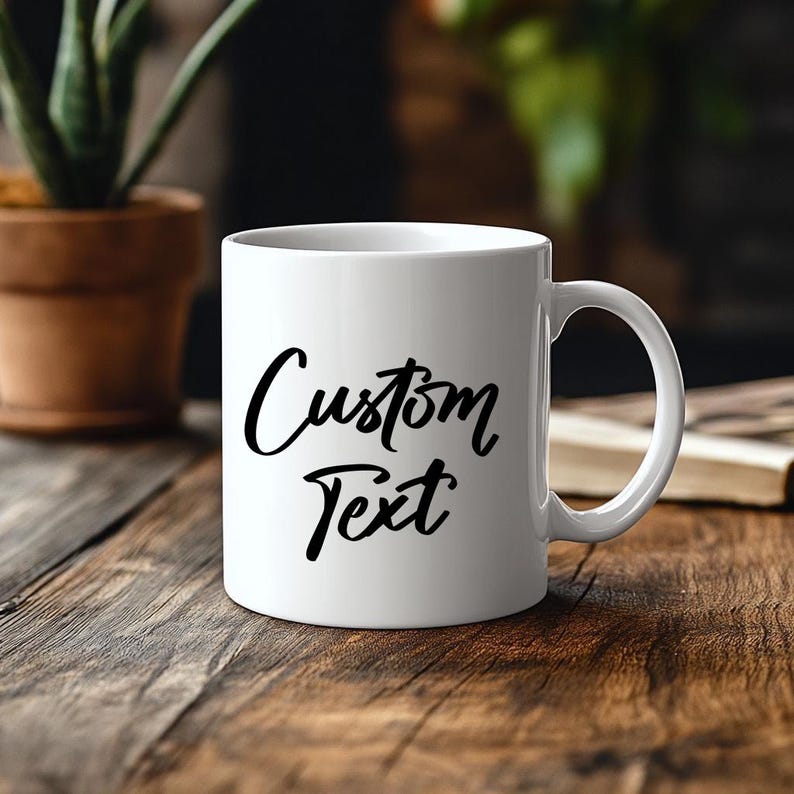 May include: A white ceramic mug with a handle on the right side, featuring the words Custom Text printed in a black, flowing, cursive-style font on the front surface. The mug has a smooth, glossy finish and a standard cylindrical shape.