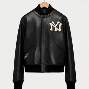 Puede incluir: Chaqueta bomber de cuero negro con cuello, puños y dobladillo de canalé negro. Cuenta con un parche con el logo "NY" color crema en el pecho y una firma en el lado izquierdo. La chaqueta tiene cierre de botones y dos bolsillos delanteros.