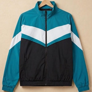 Könnte beinhalten: Türkis, weiß und schwarze Trainingsjacke mit Reißverschluss und Stehkragen. Die Jacke hat ein Zickzackmuster auf der Brust sowie einen gerippten Saum und Bündchen. Zwei Vordertaschen sind sichtbar. Die Jacke hängt an einem Holzbügel.