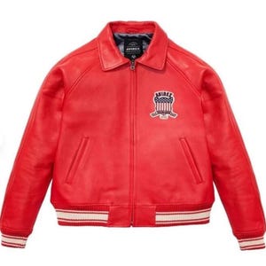 May include: A red leather jacket with a bomber-style design. The jacket features a collar, a zipper closure, and a patch on the chest with the brand name and a patriotic emblem. The cuffs and hem have white and red striped detailing.