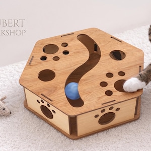 Peut inclure: Un jouet puzzle hexagonal en bois pour chat avec une finition brun clair, doté de découpes en forme de patte sur les côtés et le dessus, une piste centrale en S contenant une balle bleue et le texte JOUBERT WORKSHOP imprimé sur le dessus.