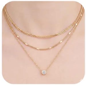 May include: A layered gold necklace with three strands. The top strand is a curb chain, the middle has small bar accents, and the bottom features a solitaire pendant with a clear gemstone. The necklace is displayed against a skin-toned background.