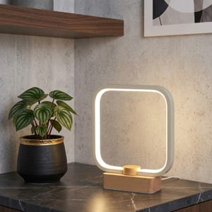 May include: A square-shaped LED table lamp featuring a white frame and a light-toned wooden base. The lamp emits a soft, warm glow from its inner perimeter. Beside the lamp sits a green-leafed plant in a black pot with a gold-coloured rim.