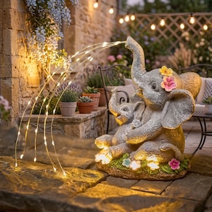 May include: A gray elephant fountain statue with a baby elephant, adorned with flowers. The fountain streams water with a string of lights. The statue is on a stone patio, with plants and string lights in the background.