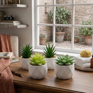 May include: Four small, artificial succulents in white geometric pots are displayed on a wooden surface. The succulents are various shades of green. A window with a brick exterior is in the background.
