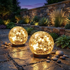 May include: Two illuminated, spherical outdoor lights with a crackled glass design. The lights are filled with warm white lights and sit on a brown base. The lights are on a stone patio with plants and a stone wall in the background.