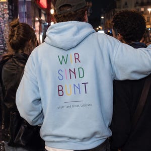 Könnte beinhalten: Ein hellblauer Kapuzenpullover mit dem Schriftzug WIR SIND BUNT in bunten Buchstaben auf der Rückseite, ergänzt durch den Text unser land bleibt tolerant, der in kleinerer Schrift unter einer horizontalen Linie im unteren Rückenbereich steht.