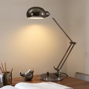 May include: A modern desk lamp with a chrome finish. The lamp features a rounded shade and an adjustable arm. A small, metallic bird figurine sits on the wooden desk, along with a pencil holder and an open book. The lamp is plugged in and turned on.