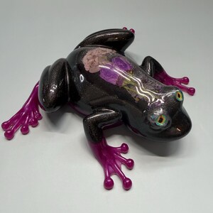 Puede incluir: Una figura decorativa de rana con un cuerpo oscuro y brillante y patas de color rosa brillante. La espalda de la rana presenta un diseño floral con flores moradas y rosas. Los ojos tienen un brillo metálico iridiscente.