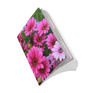 Puede incluir: Un cuaderno con una cubierta que presenta un vibrante patrón floral de numerosas flores de color rosa brillante y morado claro, similares a las margaritas, con centros oscuros. El cuaderno se muestra en ángulo, exhibiendo las hojas blancas.