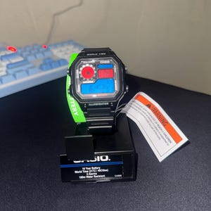 Pode incluir: Relógio digital Casio preto com mostrador retangular, com visor vermelho e azul. O relógio tem uma pulseira preta e é exibido em um suporte preto. O texto "WORLD TIME" e "ILLUMINATOR" é visível.