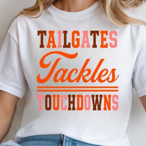 Könnte beinhalten: Ein weißes T-Shirt mit kurzen Ärmeln, das den Text TAILGATES in braunen, orangen und rosa Blockbuchstaben, das Wort Tackles in oranger Schreibschrift und das Wort TOUCHDOWNS in rosa, braunen und orangen Blockbuchstaben mit zwei orangen Linien zeigt.