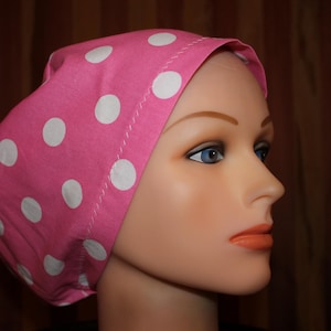 May include: A pink head covering with large white polka dots. The head covering is on a mannequin head with blue eyes and orange lipstick. The background is a dark brown.
