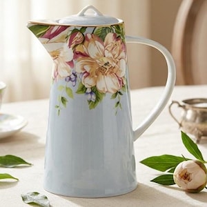 Peut inclure: Une carafe en céramique bleu clair avec un motif floral. La carafe a une poignée blanche et un couvercle. Le motif floral comprend de grandes fleurs crème et roses avec des feuilles vertes. La carafe mesure environ 25 cm de haut.