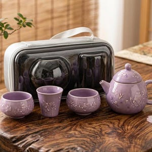Peut inclure: Un service à thé en céramique lavande comprenant une théière, trois tasses et un étui de transport. La théière et les tasses présentent des motifs floraux en relief. L'ensemble est présenté sur une surface en bois.