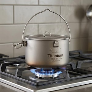 Puede incluir: Una olla de camping de titanio con asa y tapa se calienta en una estufa de gas. La olla tiene la palabra "Titanium" impresa en el lateral, junto con la capacidad de 1100 ml. Sale vapor de la olla.