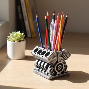 Peut inclure: Un porte-crayons argenté en forme de bloc moteur, rempli de crayons et de stylos de différentes couleurs. Une petite plante succulente en pot blanc se trouve à gauche du porte-crayons. Les crayons sont bleus, rouges, verts, violets, oranges et jaunes.