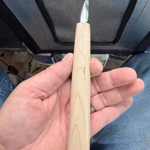 May include: A wood carving knife featuring a light-colored, smooth wooden handle with a visible grain pattern and a short, sharp, pointed metal blade extending from the top. The tool is designed for precision cutting and detailed craft work.