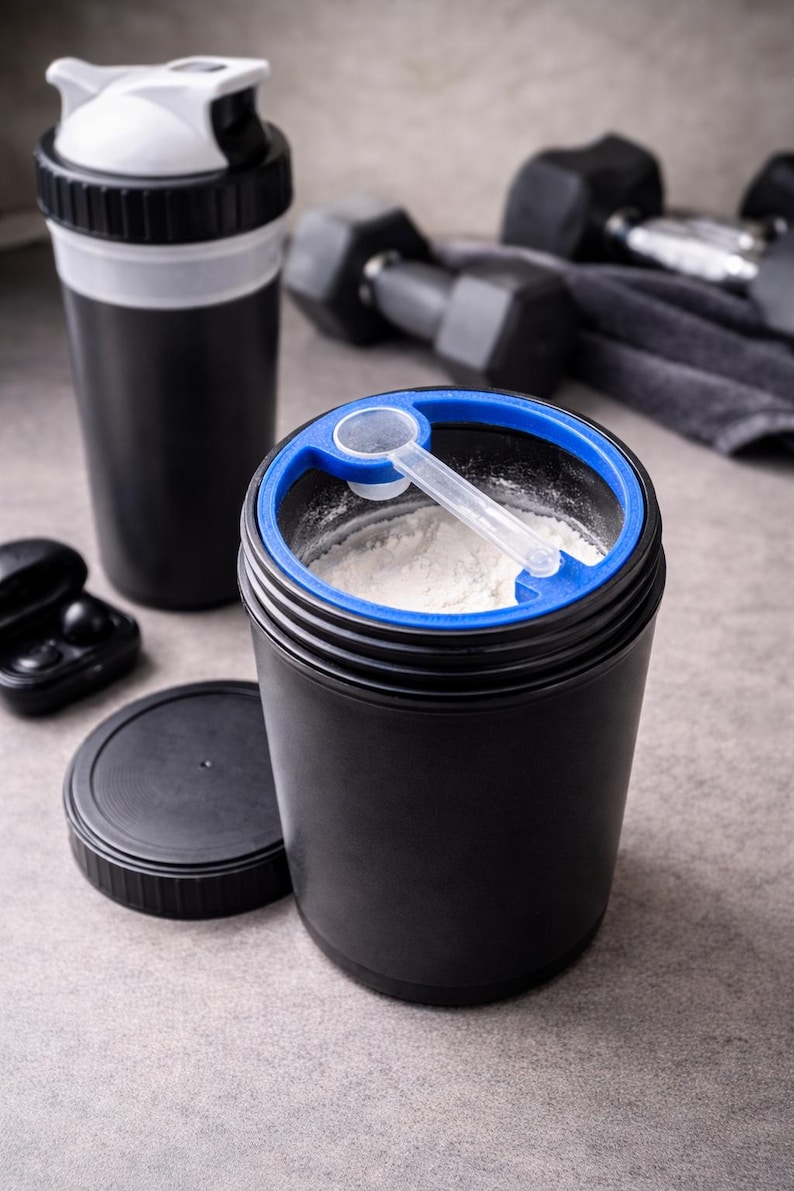 May include: A black protein powder container with a blue rim and a clear scoop, filled with white powder. A black and white shaker bottle and a pair of black earbuds are also visible. Dumbbells are in the background.