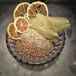 May include: A clear, faceted glass bowl containing a mixture of dried ingredients, including round brown coriander seeds, fine dried herbs, three dried orange slices, and several whole dried bay leaves arranged on top.