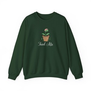May include: A dark forest green crewneck sweatshirt featuring a central graphic of a pink and green Venus flytrap plant in a brown terracotta pot. Below the plant, the text "Feed Me" is printed in a white, elegant, cursive script font.