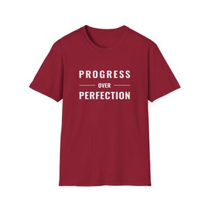 May include: A maroon t-shirt featuring the white text "PROGRESS OVER PERFECTION". The text is stacked with a line separating the two words. The shirt has a crew neck and short sleeves.