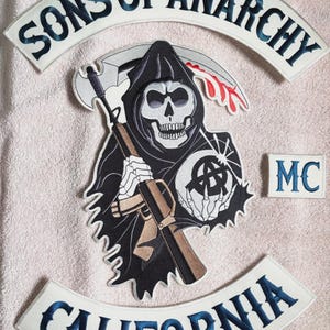 May include: A set of embroidered patches featuring a central grim reaper figure holding a rifle and a scythe, with the text SONS OF ANARCHY in a top arch, CALIFORNIA in a bottom arch, and a small rectangular patch with the letters MC.