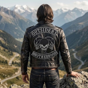 May include: A black leather jacket featuring a white embroidered patch on the back. The patch displays three skulls and the text OUTSIDERS BROTHERHOOD in a curved font. The jacket has long sleeves with zippered cuffs and a structured collar.