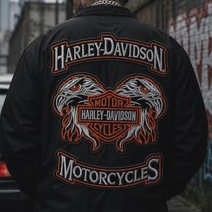 Könnte beinhalten: Eine schwarze Jacke mit einem großen gestickten Rückenaufnäher mit dem Schriftzug Harley-Davidson Motorcycles. Das Design zeigt zwei Adlerköpfe, die ein zentrales orange-schwarzes Harley-Davidson Motor Cycles Logo mit weißen Akzenten flankieren.