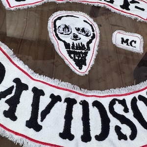 Könnte beinhalten: Ein Set aus drei bestickten Stoffaufnähern mit weißem Hintergrund, schwarzer Schrift und rotem Rand. Der obere geschwungene Aufnäher zeigt HARLEY-, der untere geschwungene DAVIDSON und ein kleinerer rechteckiger Aufnäher trägt die Aufschrift MC.