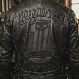 Op de afbeelding: Een zwart leren jack met een verweerde uitstraling, voorzien van een grote rugpatch met de tekst Harley-Davidson bovenaan, een nummer één-afbeelding met een schedelontwerp in het midden en het woord Motorcycles onderaan op een gebogen patch.