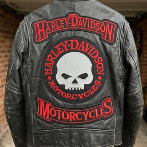 Puede incluir: Una chaqueta de cuero negro con un gran parche bordado en la espalda. El parche muestra un gráfico central de una calavera gris claro rodeado por texto rojo que dice Harley-Davidson Motorcycles, con texto adicional de Harley-Davidson arriba y abajo.