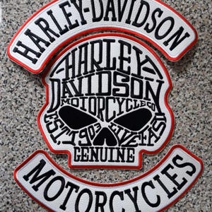 Könnte beinhalten: Ein dreiteiliges besticktes Aufnäher-Set in den Farben Weiß, Schwarz und Rot. Die oberen und unteren Bogenaufnäher tragen die Schriftzüge Harley-Davidson und Motorcycles. Der mittlere Aufnäher in Totenkopfform besteht aus dem Text Harley-Davidson Motorcycle Co Est 1903.