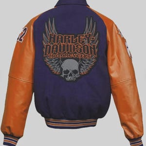 Könnte beinhalten: Eine violette und orangefarbene Collegejacke mit einer großen Stickerei auf der Rückseite, die den Schriftzug Harley-Davidson Motorcycles über einem Totenkopf mit Flügeln zeigt. Die Ärmel sind aus orangefarbenem Leder, Kragen und Bündchen gestreift.
